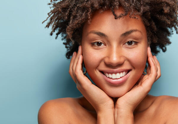 Close Up Portrait Of Relaxed Black Woman Has Gentle Skin After Taking Shower, Satisfied With New Lotion, Has No Makeup, Smiles Tenderly, Shows Perfect Teeth, Stands Shirtless Against Blue Background